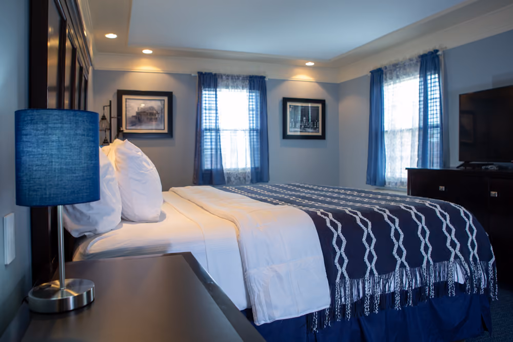 Side view of king bed with white linens and navy throw in a guest room with two large windows and television