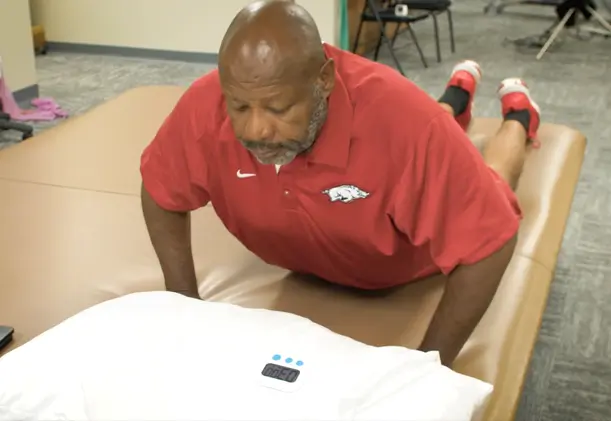 A man performs a McKenzie Method (MDT) prone press-up exercise on a treatment table, using his arms to raise his upper body while keeping his hips and legs supported.