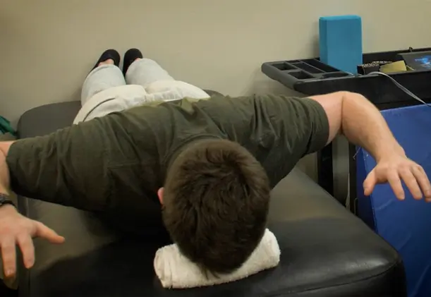 A man lies face down on a treatment table with his arms extended out to the sides while performing spinal stabilization exercises.