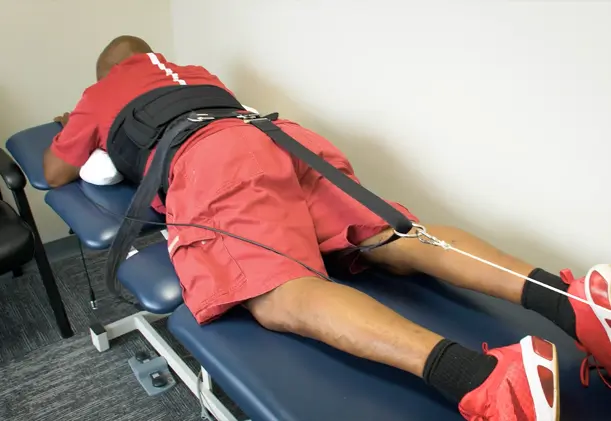 A man lies face down on a padded treatment table while receiving non-surgical spinal decompression therapy, with a harness secured around his lower back and pelvis and connected to a traction system.