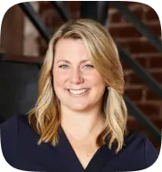 Smiling blonde woman with shoulder-length hair wearing a dark blue top, with a blurred brick wall background.