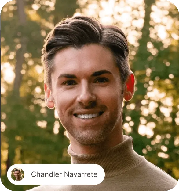Smiling man with neatly styled dark hair wearing a brown turtleneck sweater outdoors with blurred green foliage background.