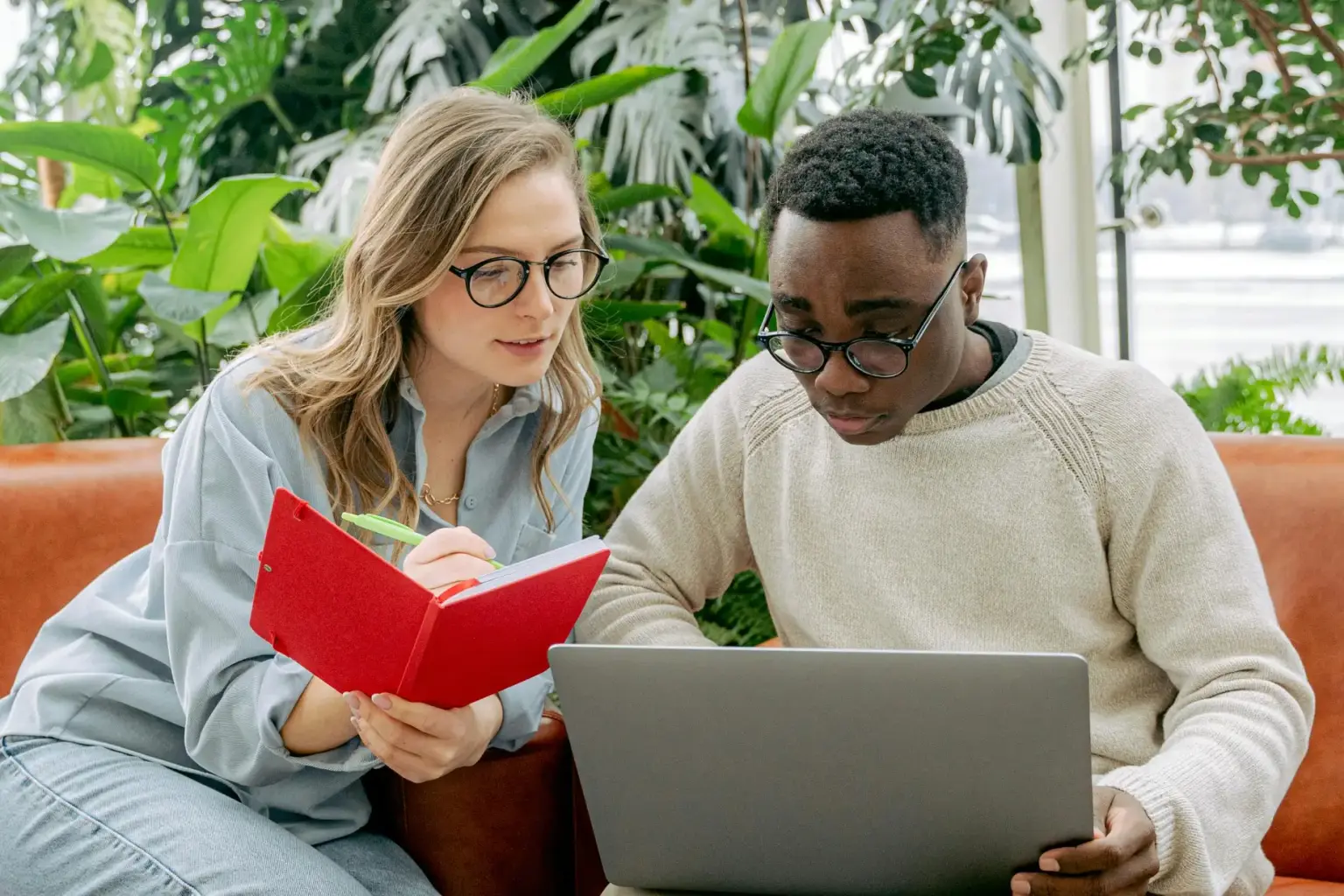 A woman with glasses writing in a red notebook while a man with glasses looks at a laptop screen in a green plant-filled room.