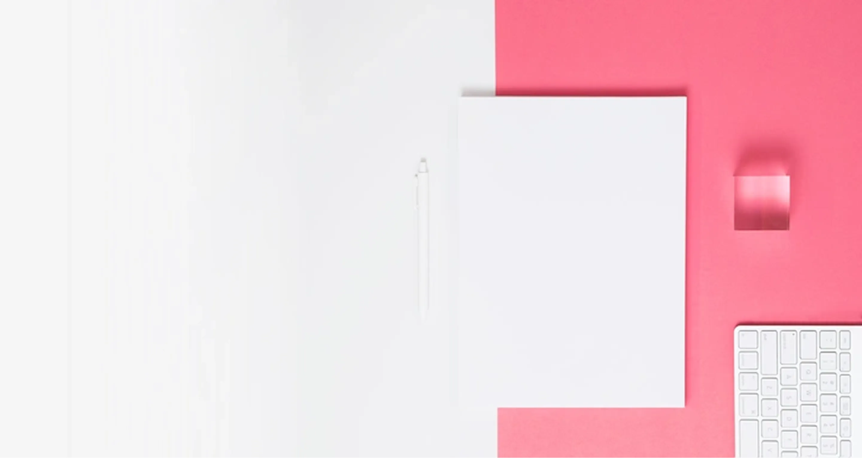 Top view of a white paper sheet, white pen, transparent cube, and white keyboard on a pink and white background.