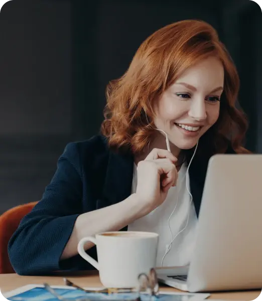 Smiling red-haired woman with earphones working on a laptop with a white coffee cup in front of her.