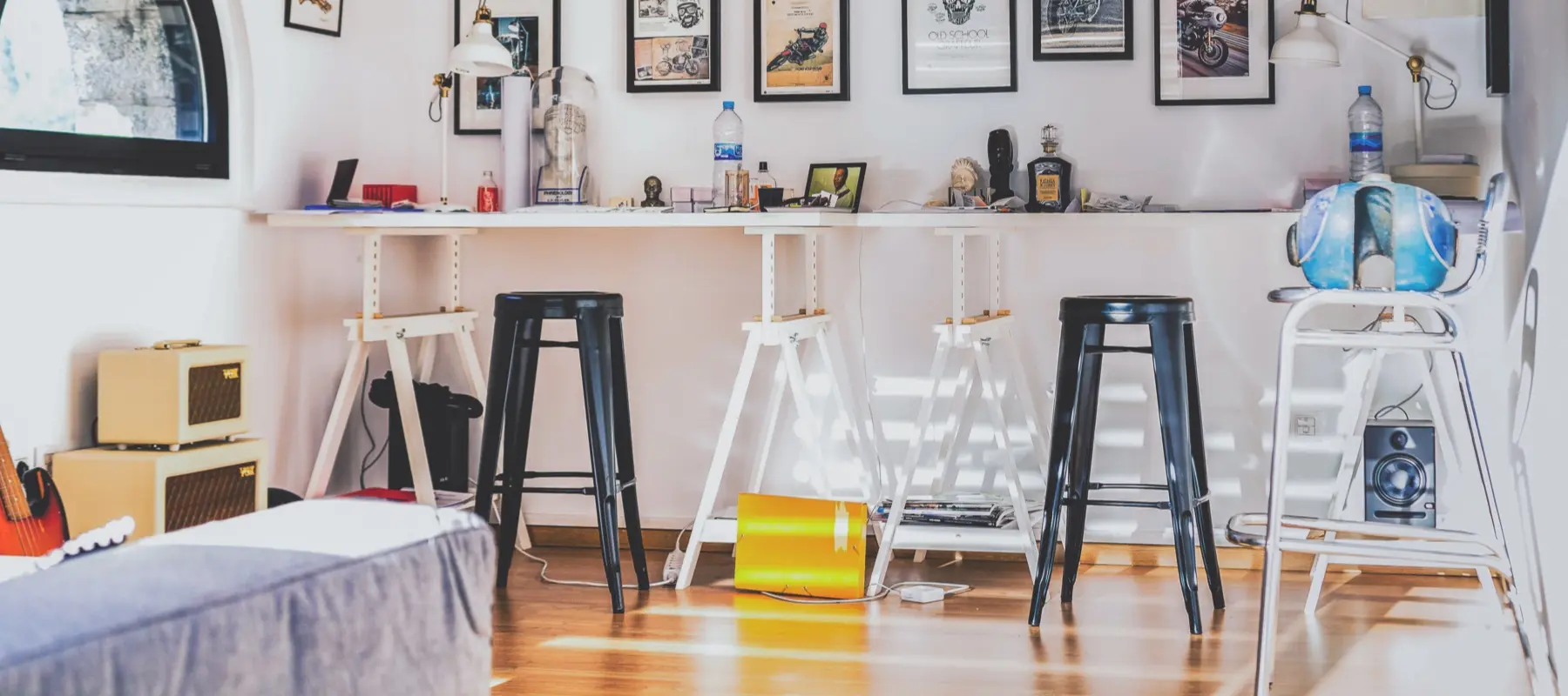 Bright room with stools, a white desk, art on the wall, guitars, and speakers on wooden floor.