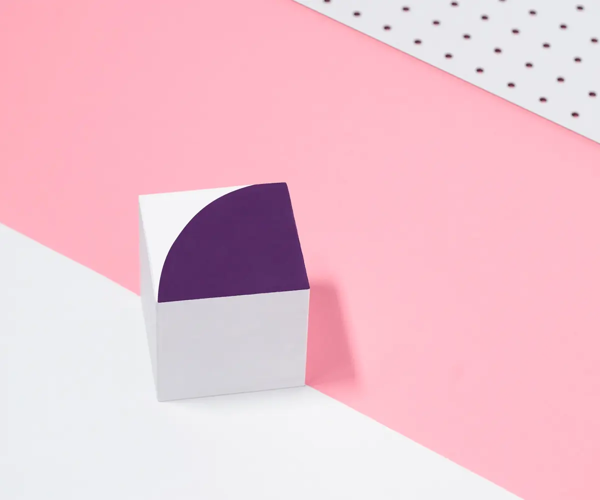 White cube with a dark purple quarter circle on top, on a pink and white background with polka dots.
