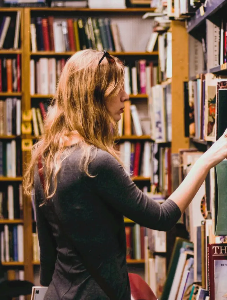 Woman with long hair browsing books on a shelf in a bookstore or library.