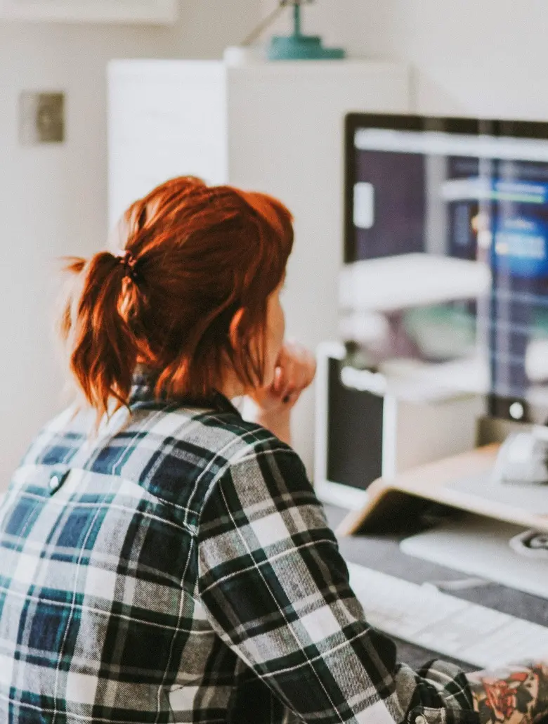 Woman with red hair in a plaid shirt sitting at a desk, looking at a computer monitor.