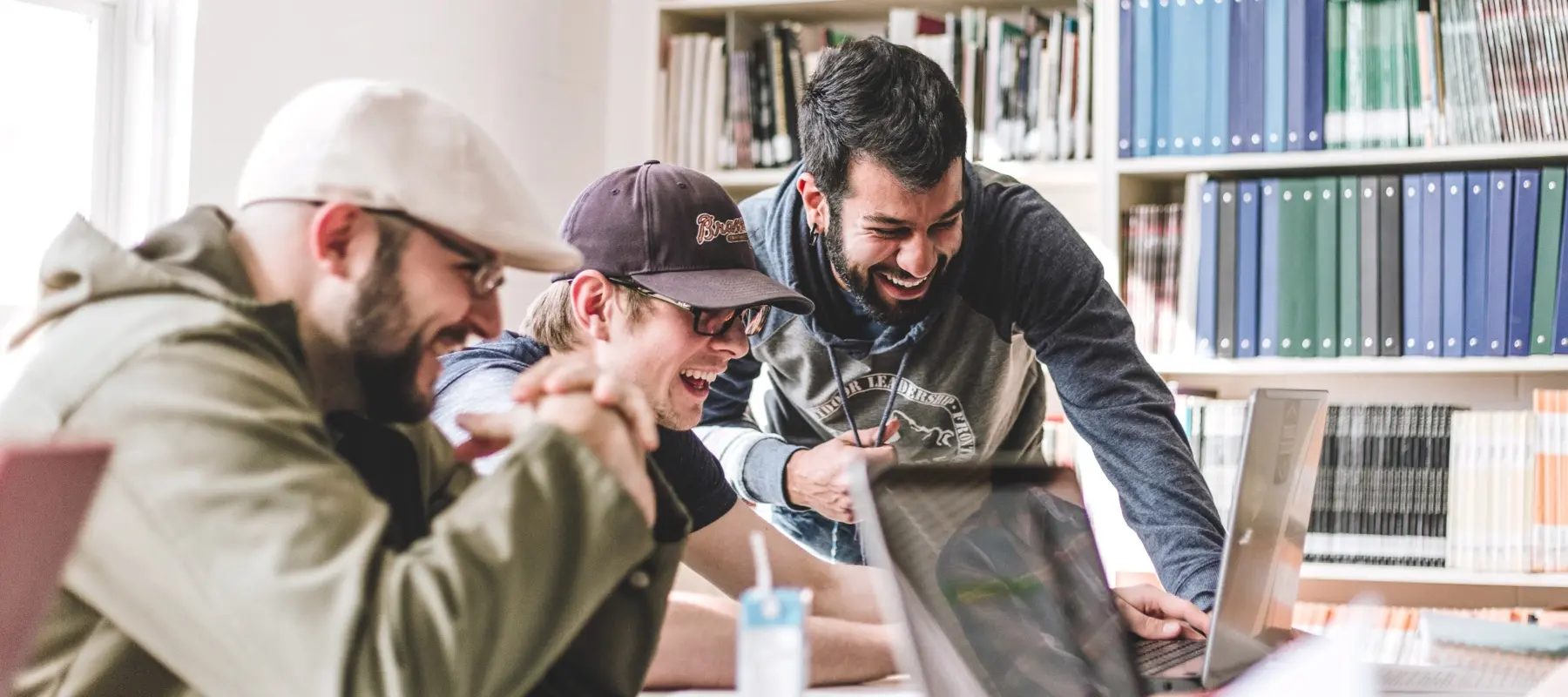 Three young men smiling and laughing while looking at laptops in a bright room with bookshelves.