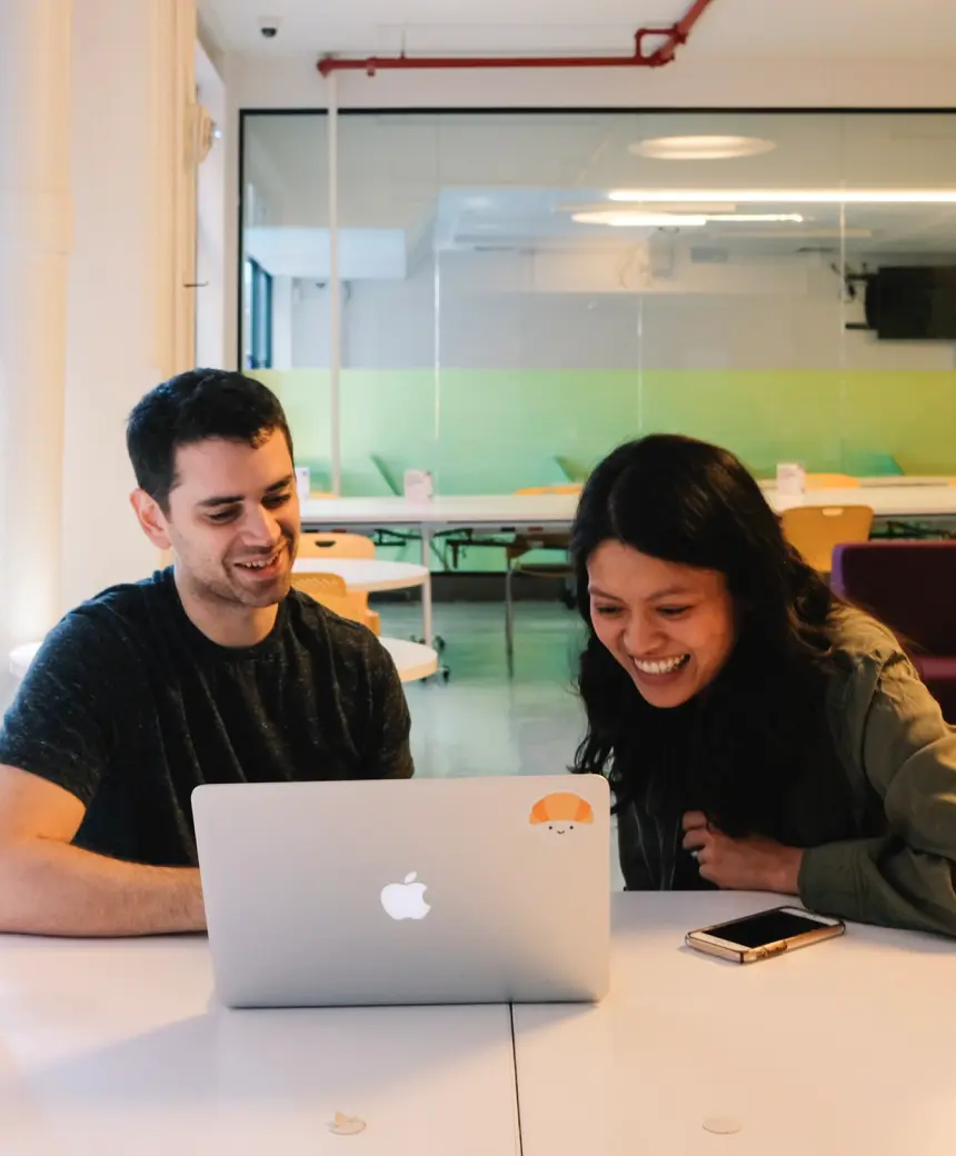Two people smiling and looking at a laptop with a croissant sticker, sitting in a modern office.