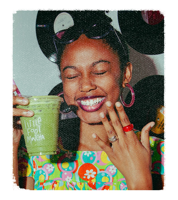 Zariah in a stamp. Smiling with her eyes closed, showing off a manicure. Holding a green smoothie.