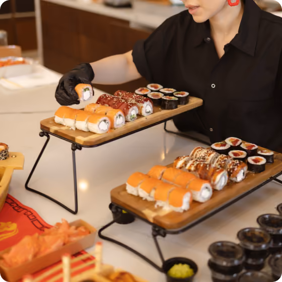 Person wearing black glove picking sushi roll from wooden tray with assorted sushi on table.