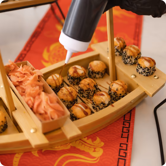 Sauce being drizzled on sushi balls with black sesame seeds arranged on a wooden serving boat.