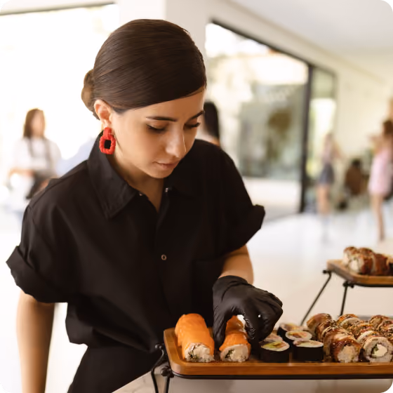 Woman in black shirt and gloves arranging sushi rolls on a wooden serving tray.