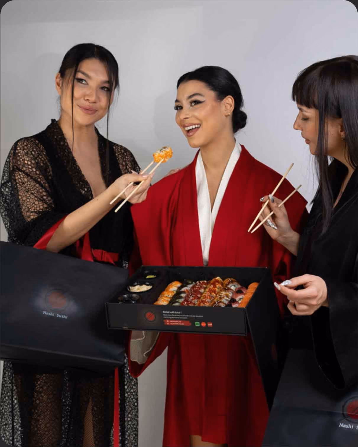 Three women in robes holding chopsticks and a box of assorted sushi rolls, smiling together.