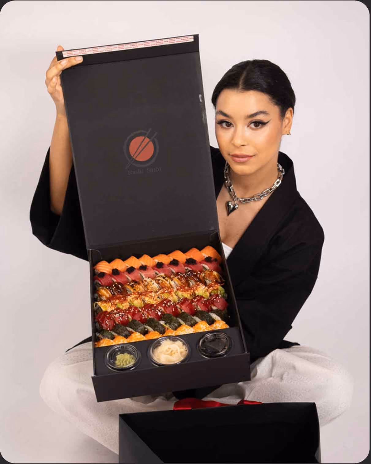 Woman holding open large box with assorted sushi rolls and condiments inside.