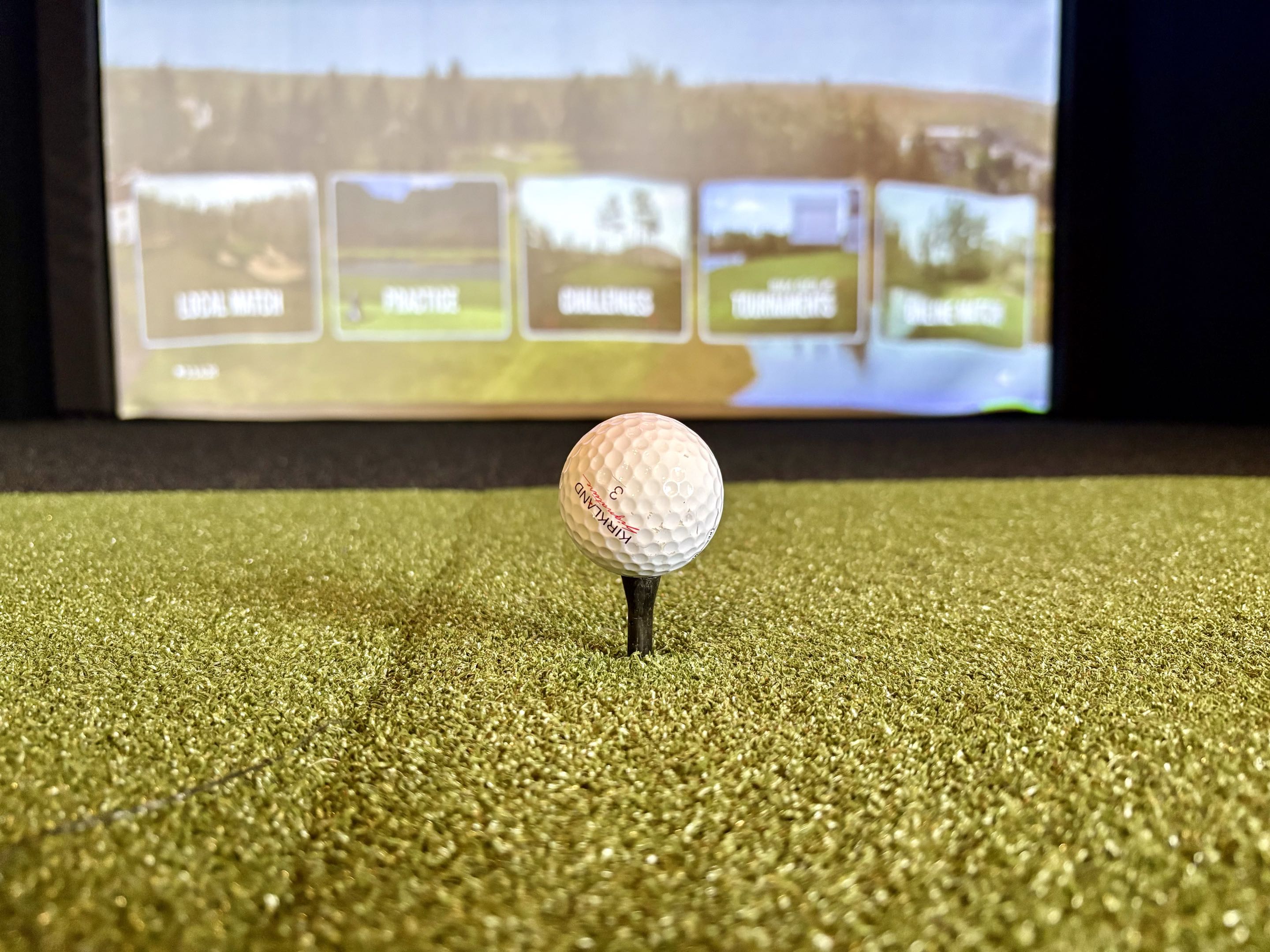 Golf ball on a tee inside a GridZone indoor golf simulator bay with a virtual course displayed on the screen.