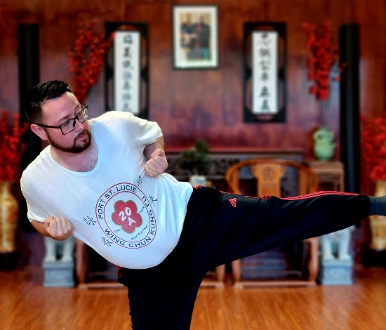 Man wearing glasses performing a martial arts side kick in a room with wooden floor and Asian decor.