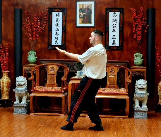 Man in a martial arts stance in a room with wooden chairs, statues, and framed calligraphy.