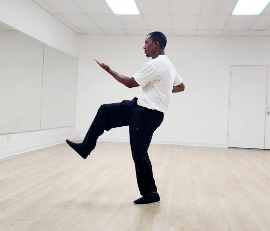 Man in white shirt and black pants balancing on one leg with arm extended in a dance studio.