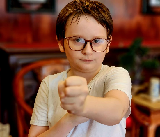 Boy with glasses in white shirt showing a fist forward indoors with a blurred background.