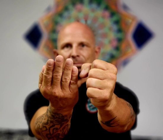 Man with tattoos holding hands forward, one open palm and one fist, blurred patterned background.