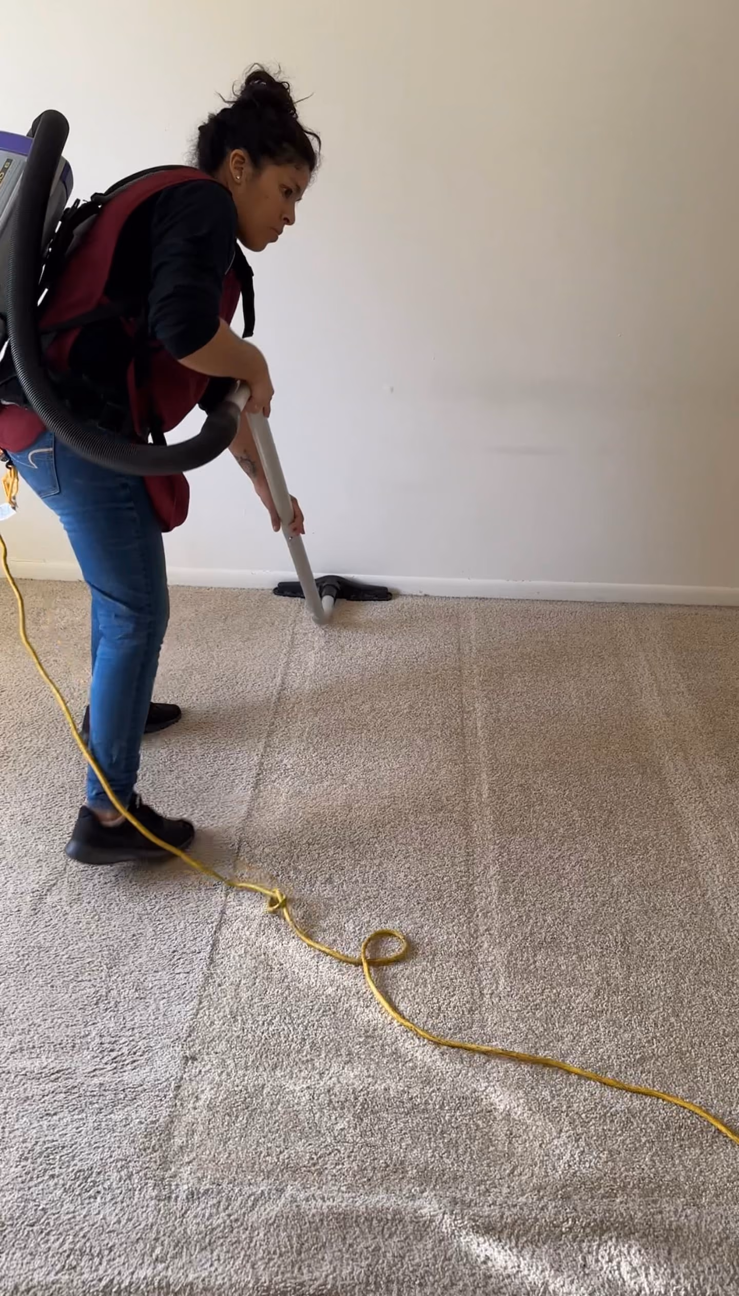 Cleaner using a backpack vacuum on beige carpet in an empty room.
