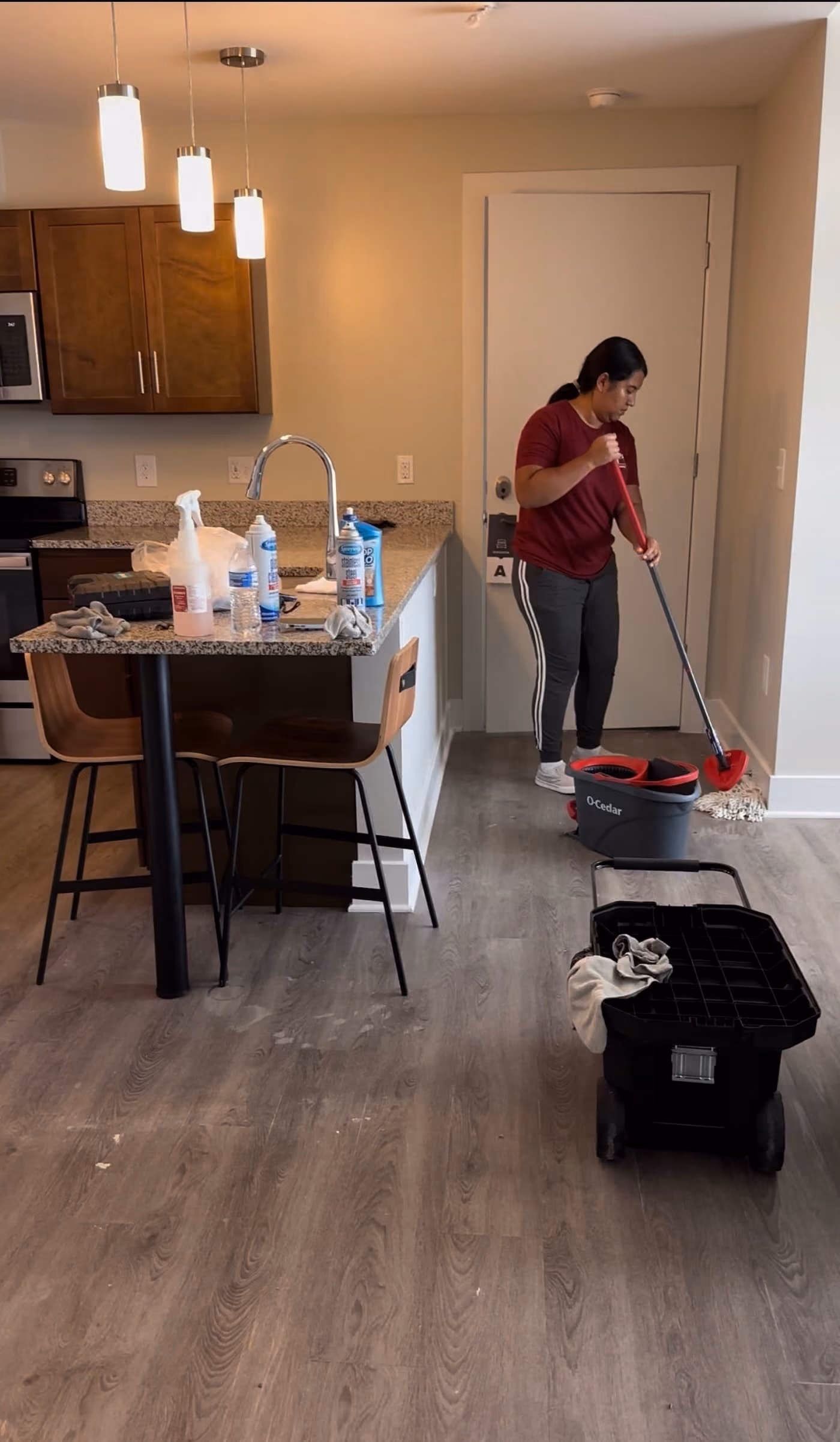 Cleaner mopping the floor of an open-plan apartment kitchen with a cleaning cart nearby.