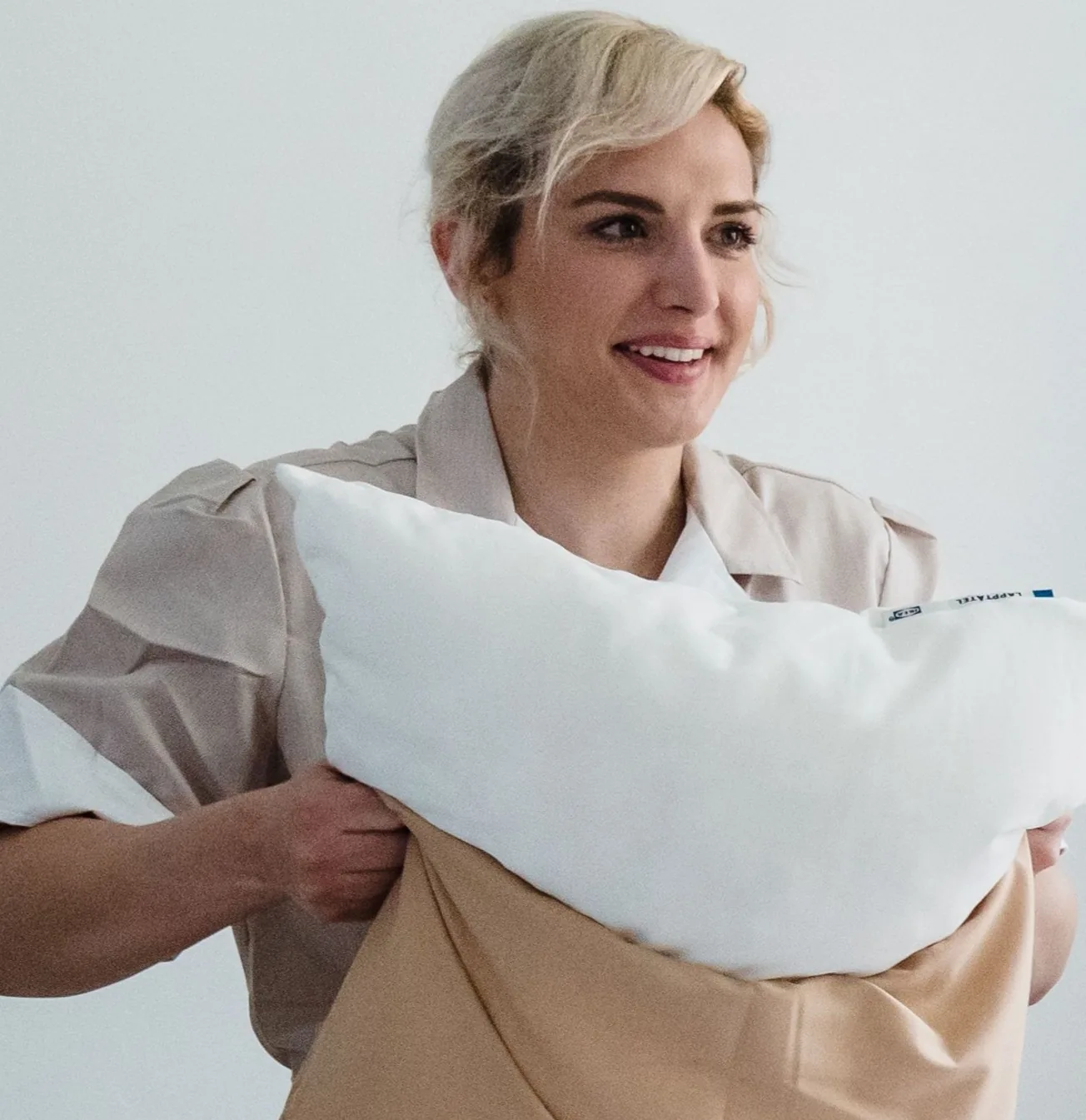 Smiling cleaner holding a fluffed white pillow while making a bed.