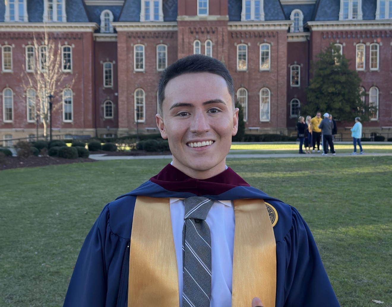 Patrick Long smiling in graduation regalia on a college campus lawn.