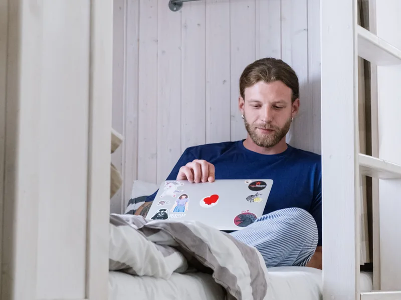 Young man sitting on a bed working on a laptop covered in stickers.