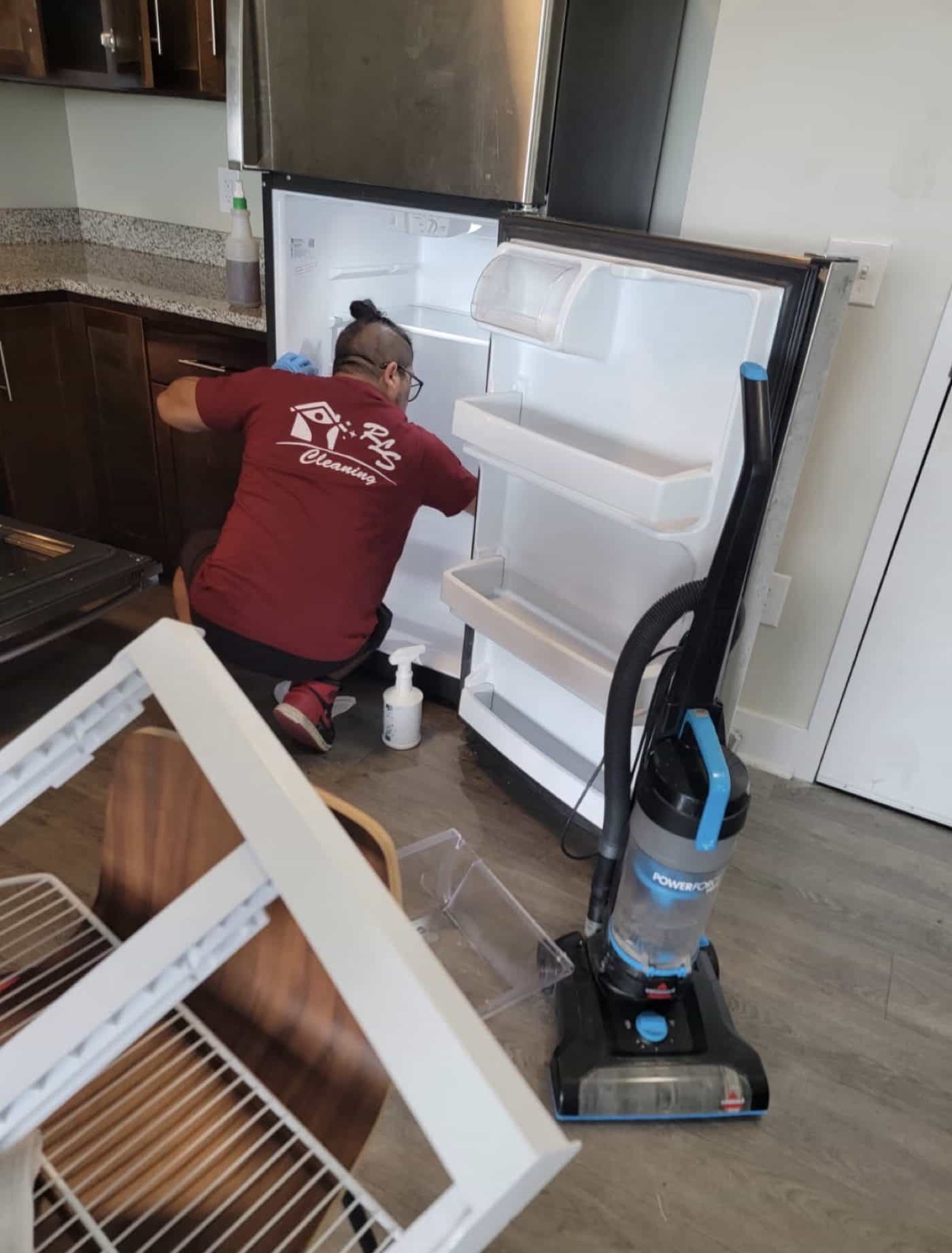 Cleaner kneeling at an open, empty refrigerator with its shelves and drawers pulled out.