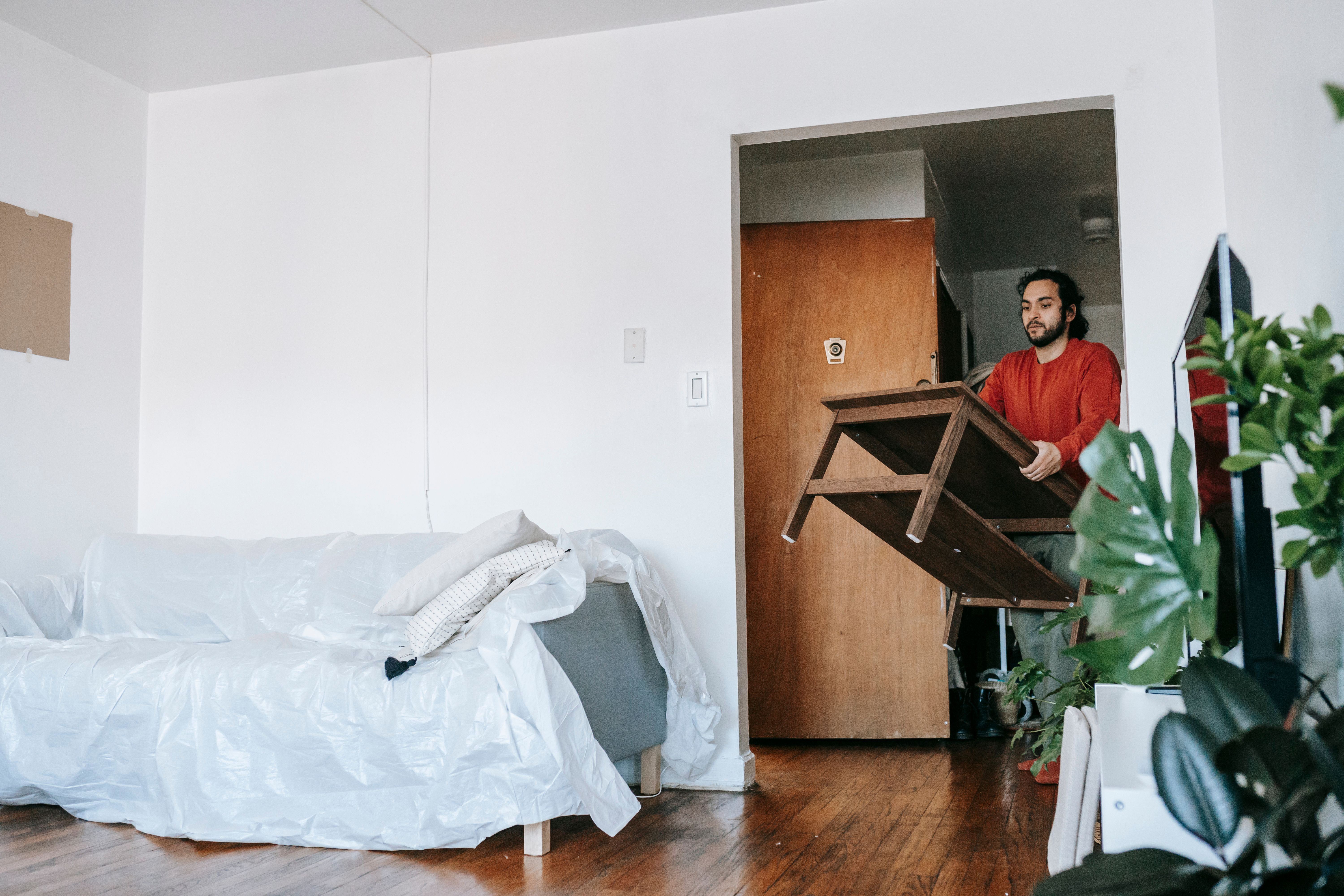  Tenant carrying a wooden table out of an apartment with the sofa covered in plastic on move-out day