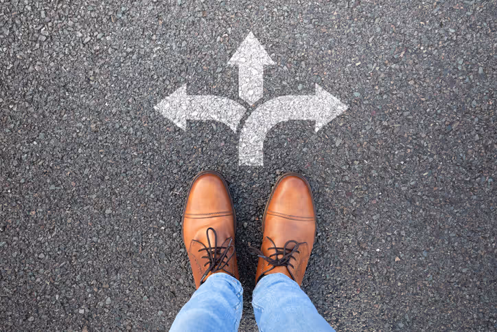 Looking down at a person's shoes on asphalt, with three painted white arrows pointing left, straight ahead and right