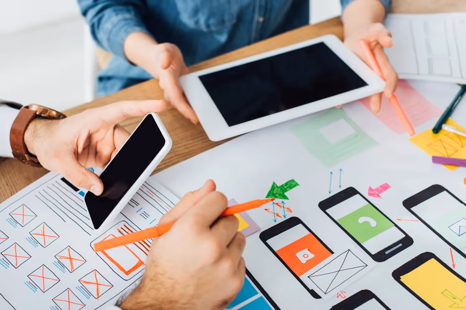 Two colleagues at a desk reviewing printed mobile app screen layouts, holding a tablet and a smartphone