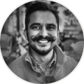 Smiling man with a mustache and beard, wearing a buttoned shirt in an outdoor setting.