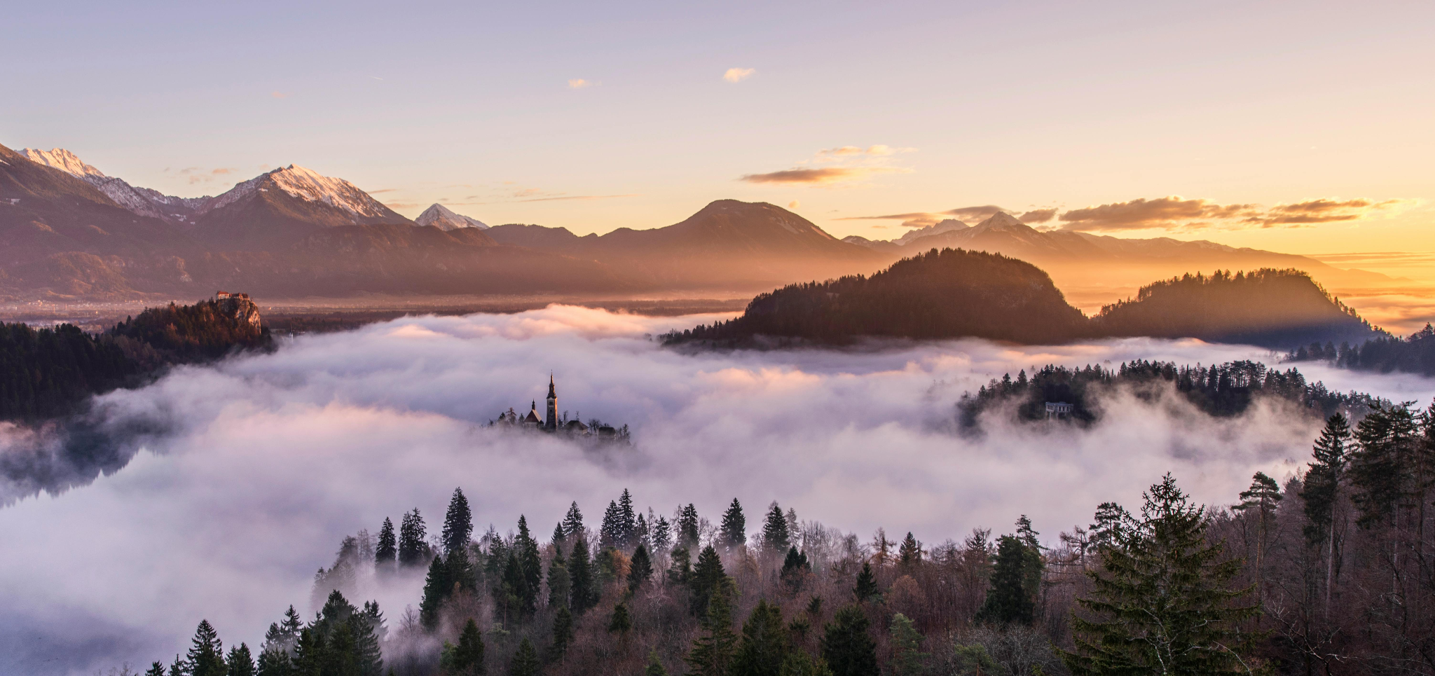 Fog covered landscape with church steeple, forested hills, and snow-capped mountains at sunrise.