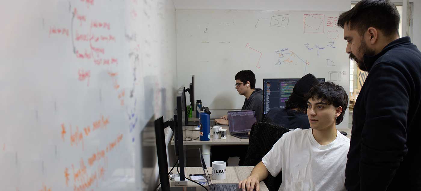 Four people working on computers in an office with whiteboards covered in diagrams and notes.