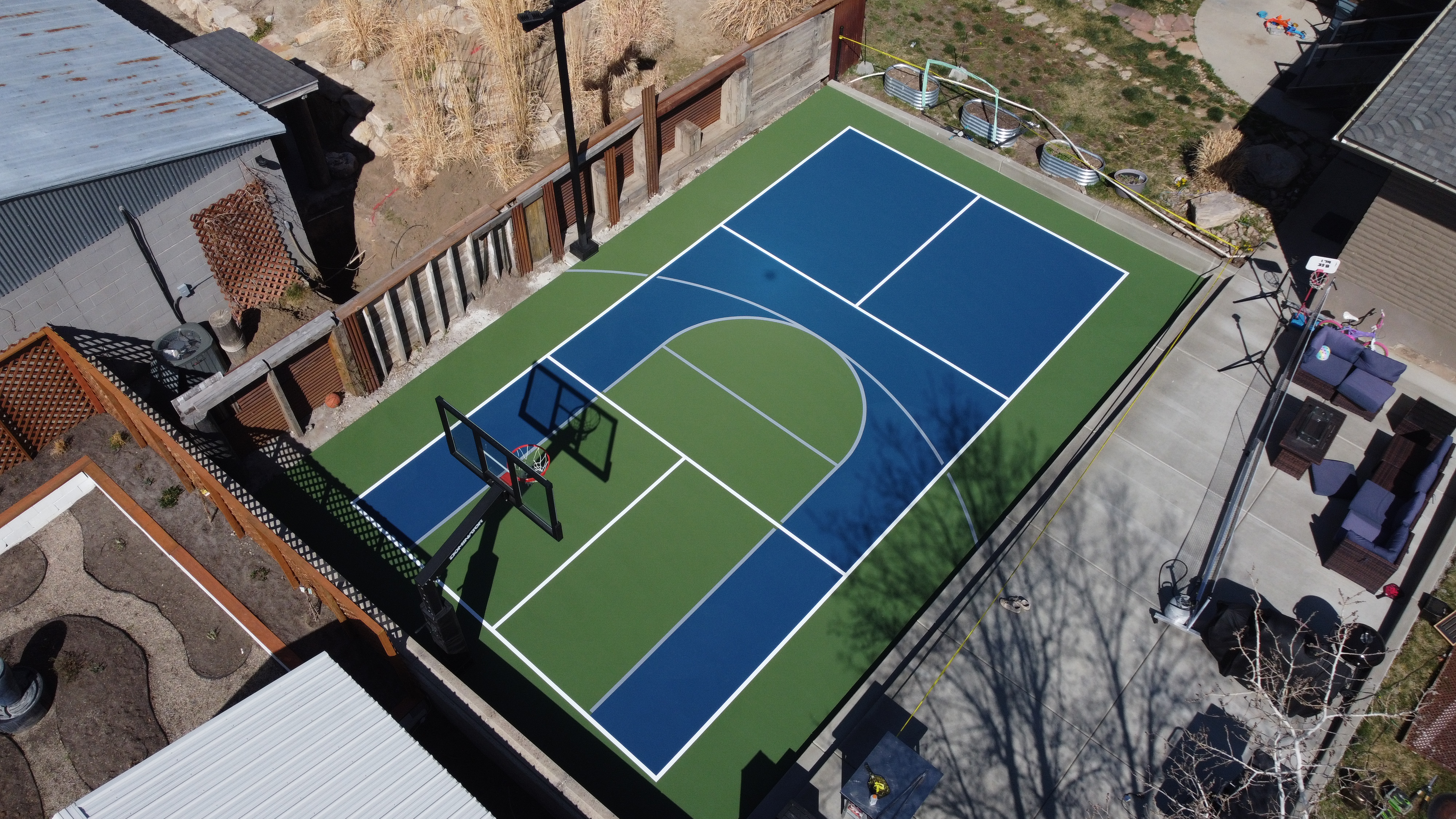 Aerial view of a backyard sports court with basketball and tennis markings surrounding outdoor furniture.