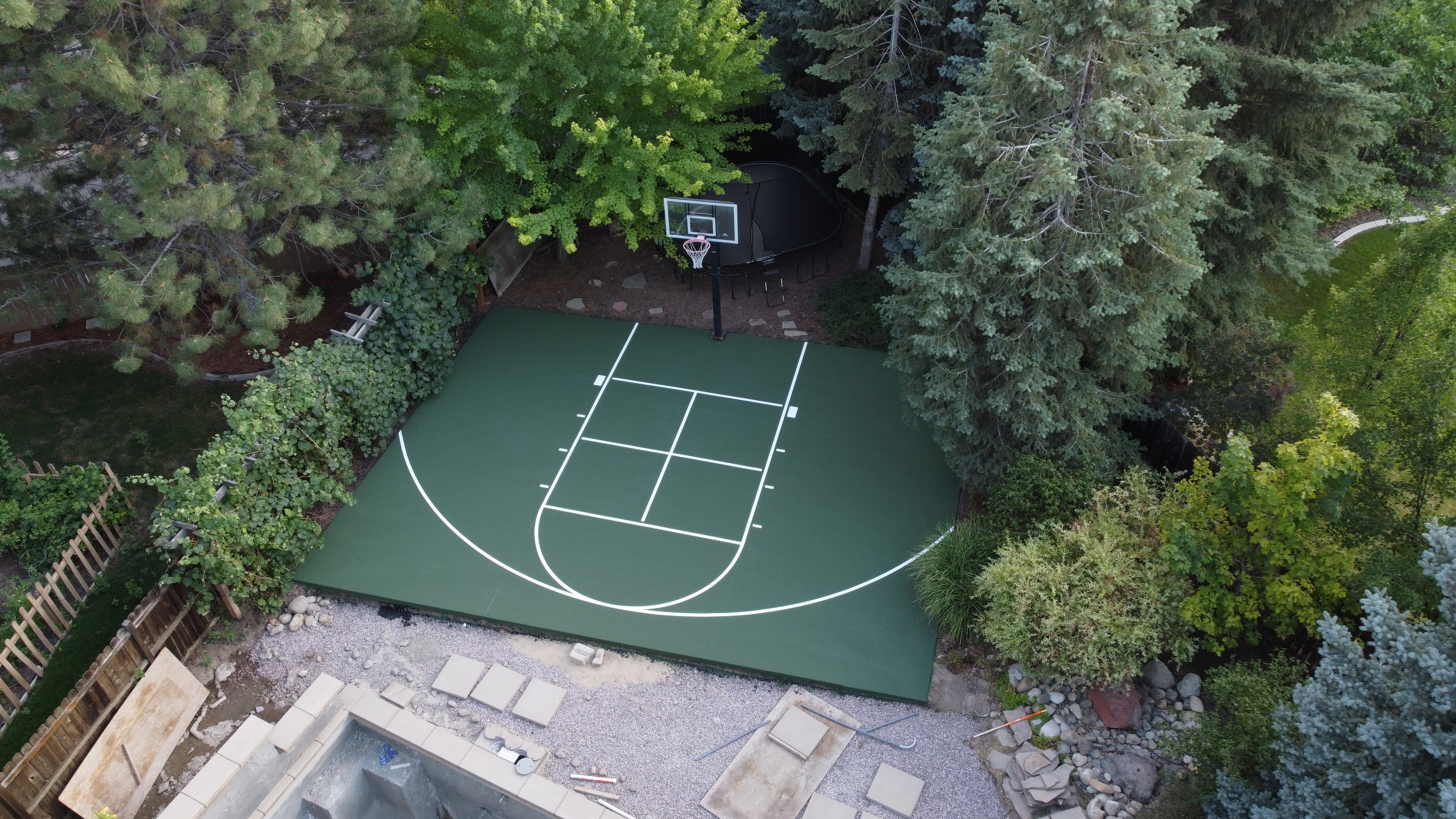 A small green outdoor basketball court surrounded by dense trees and shrubs in a backyard.