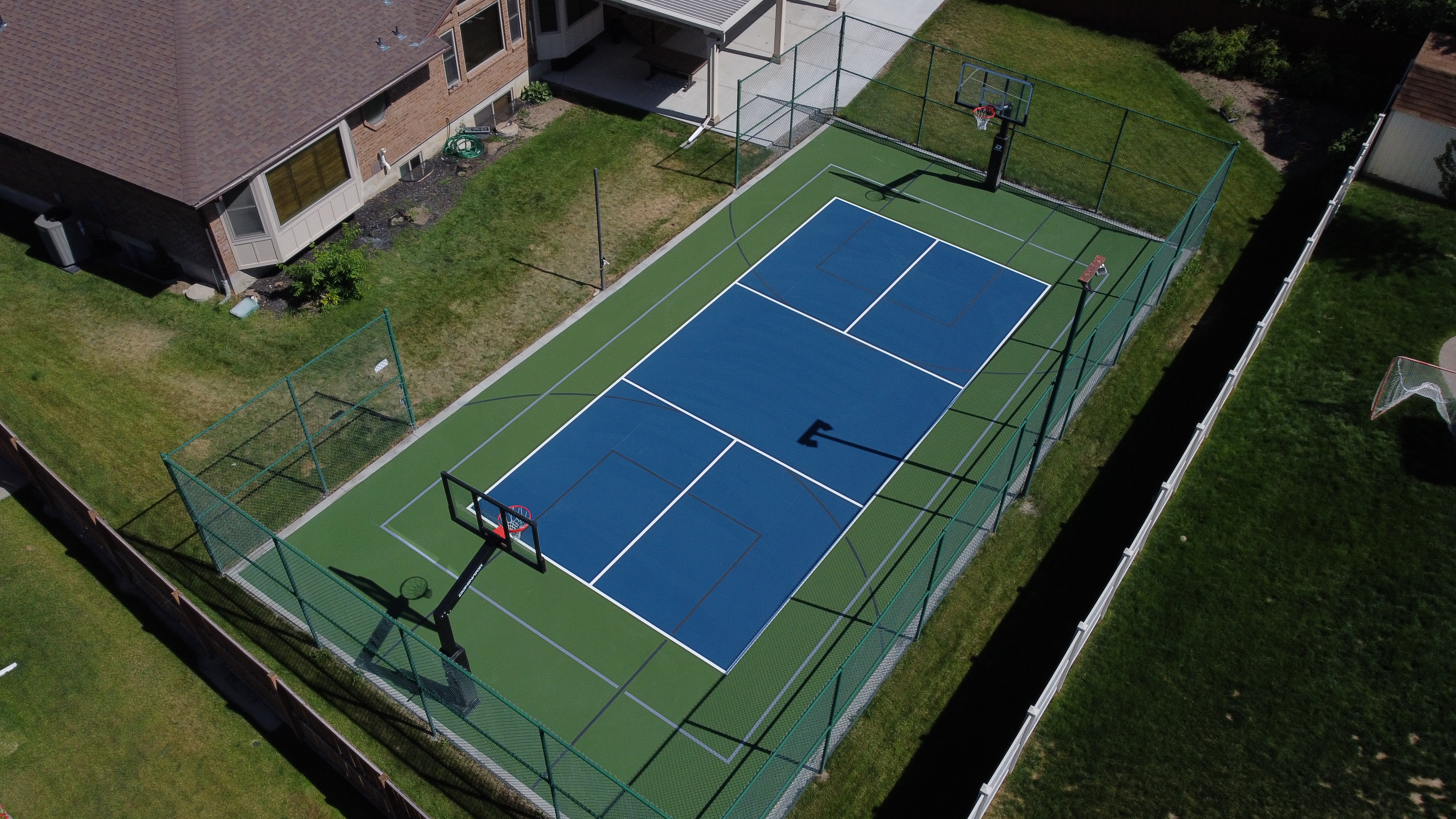 A fenced blue and green private tennis and basketball court beside a brick house in a grassy yard.