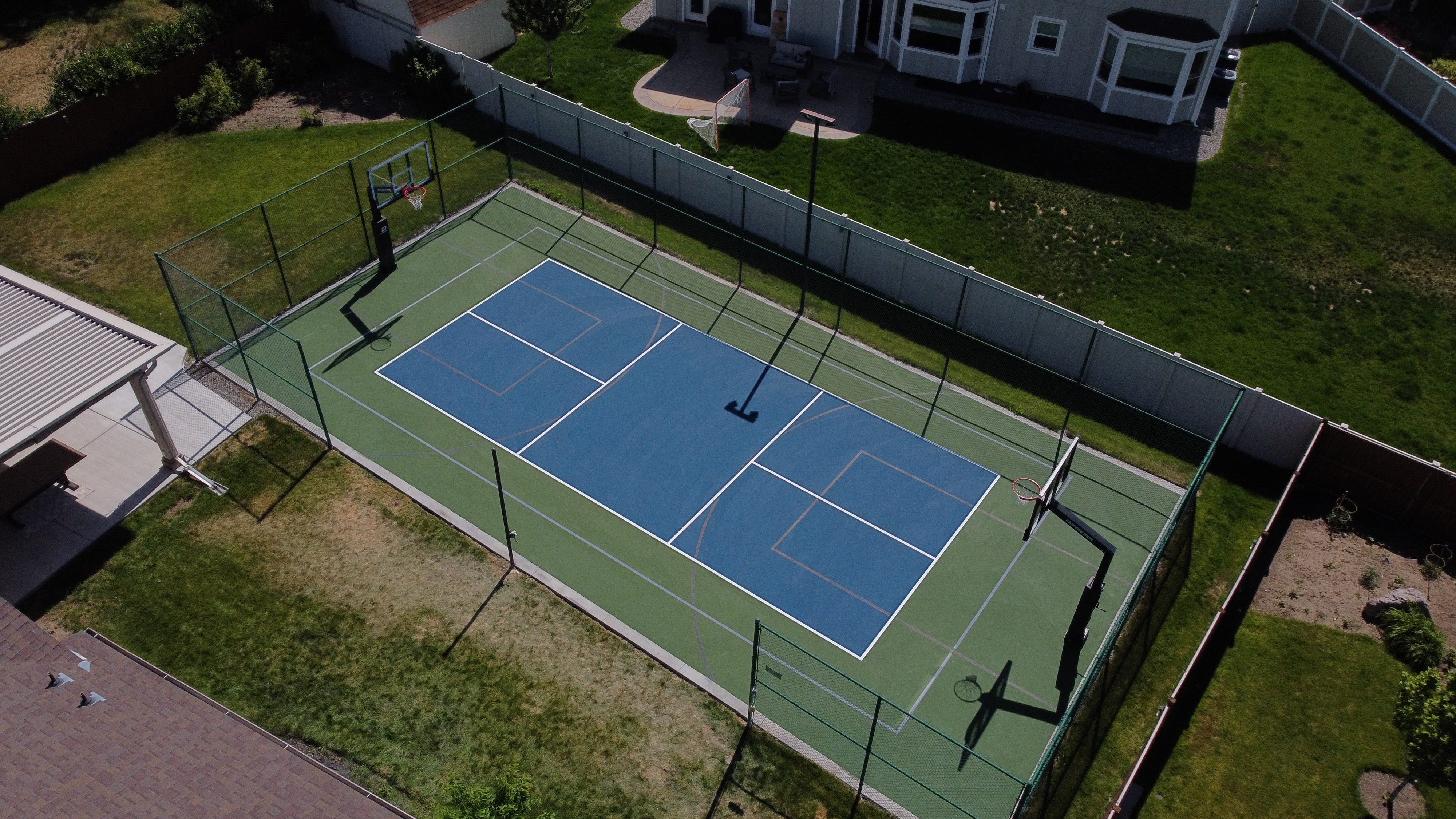 A backyard sports court with basketball hoops and a fenced tennis or pickleball court under daylight.