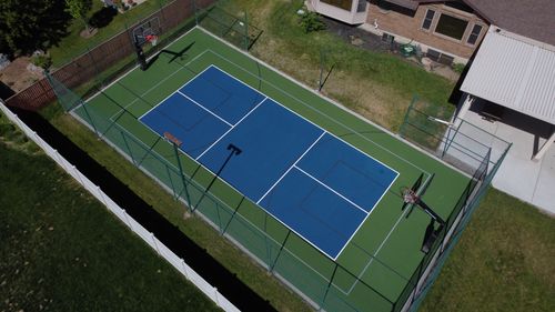 A fenced, blue and green outdoor basketball court next to a house and lawn.