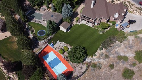 Aerial view of large backyard with tennis court, trampoline, fire pit area, and lawn near a house.