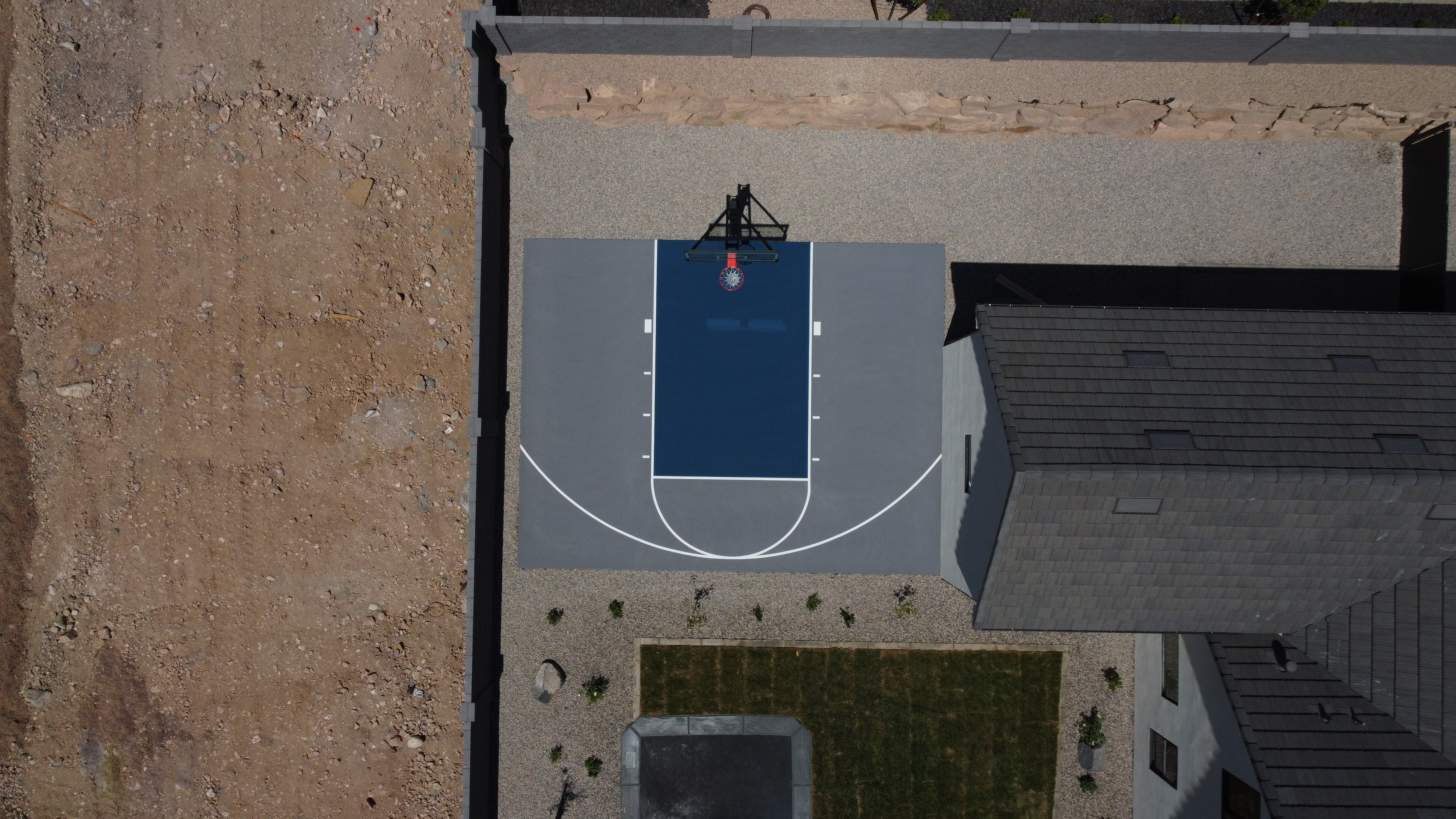 Aerial view of a small basketball half-court next to a modern house with gravel and grass landscaping.