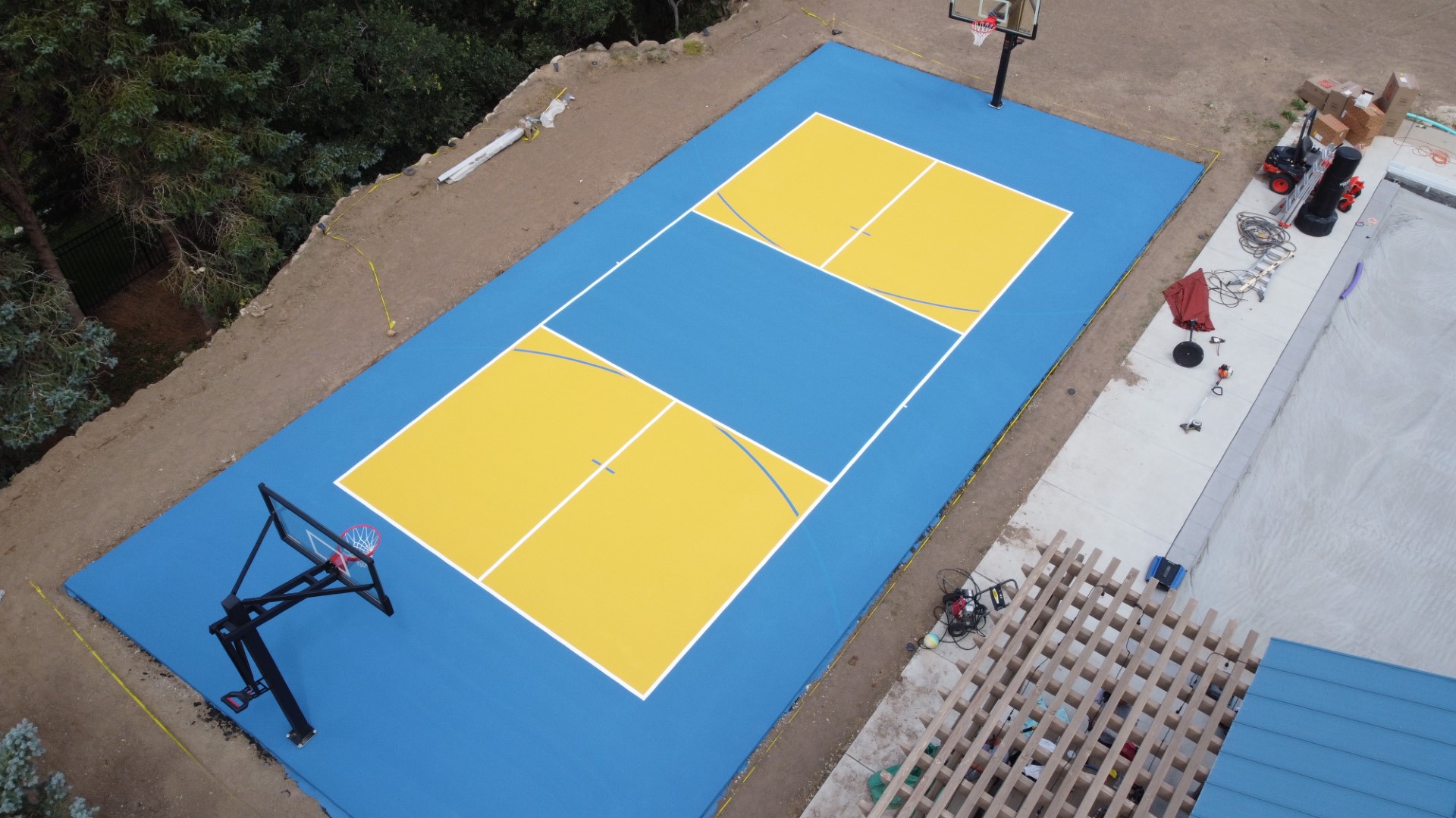 Aerial view of a blue and yellow outdoor basketball court with two hoops surrounded by dirt and trees.