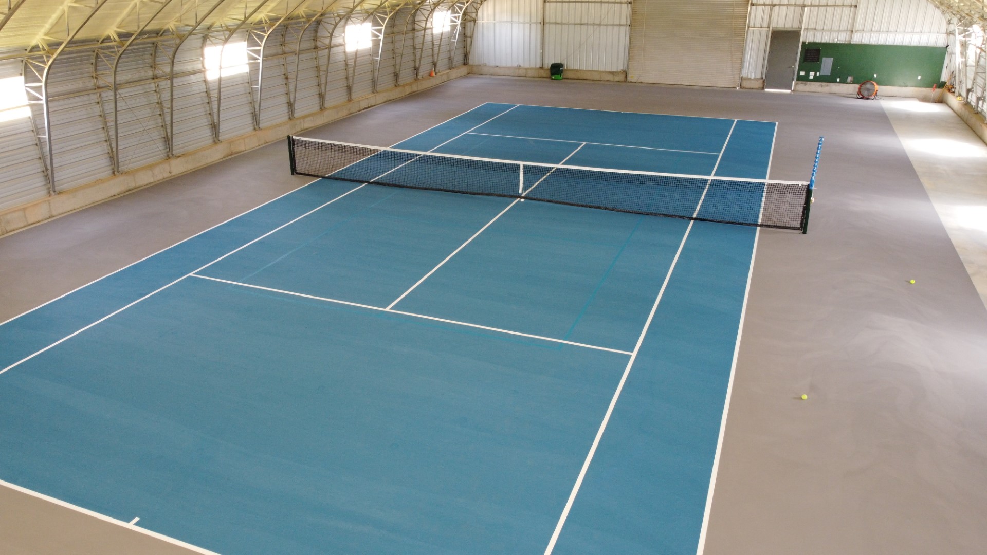 Indoor tennis court with blue playing surface, white lines, black net, and two tennis balls on the floor.