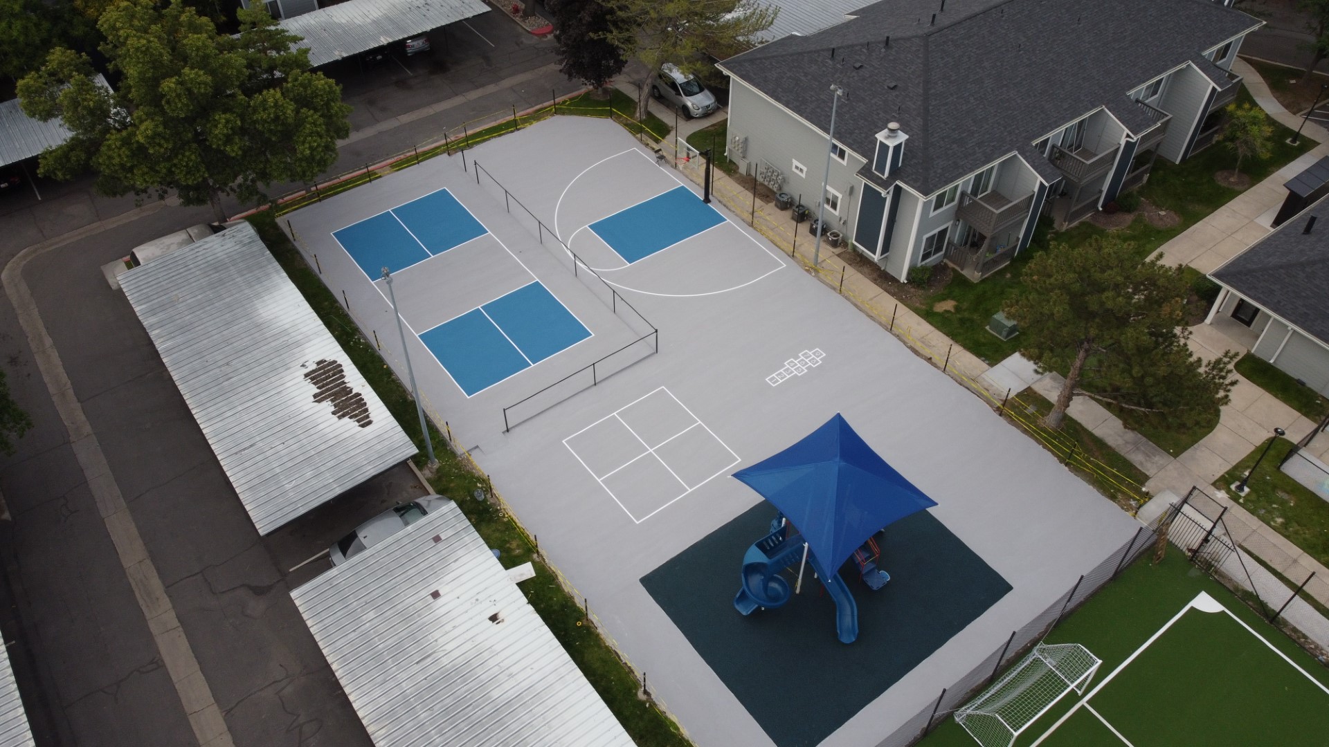 Aerial view of a playground with a blue slide, basketball court, pickleball courts, and hopscotch grid.