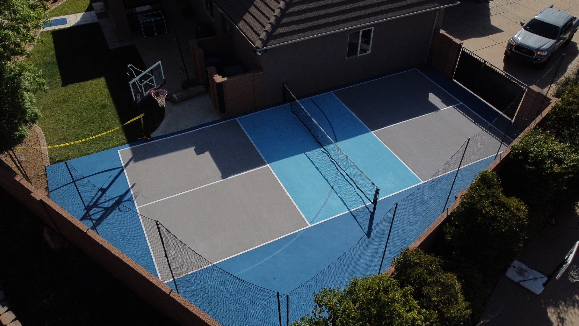 Backyard sports court with a basketball hoop and a net for tennis or pickleball next to a house.